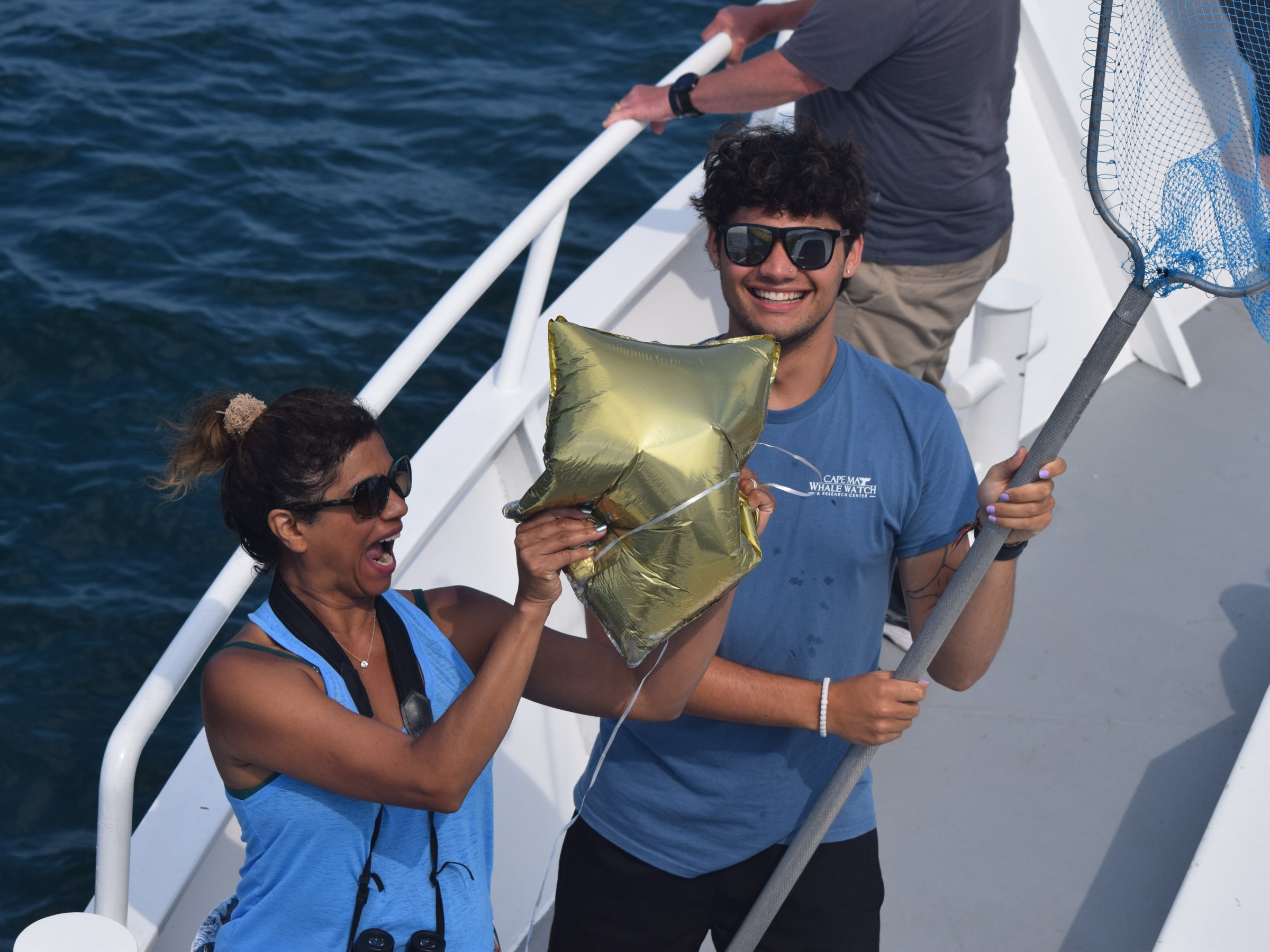 Two people on a boat holding a golden star-shaped balloon and a net, smiling with the ocean in the background.