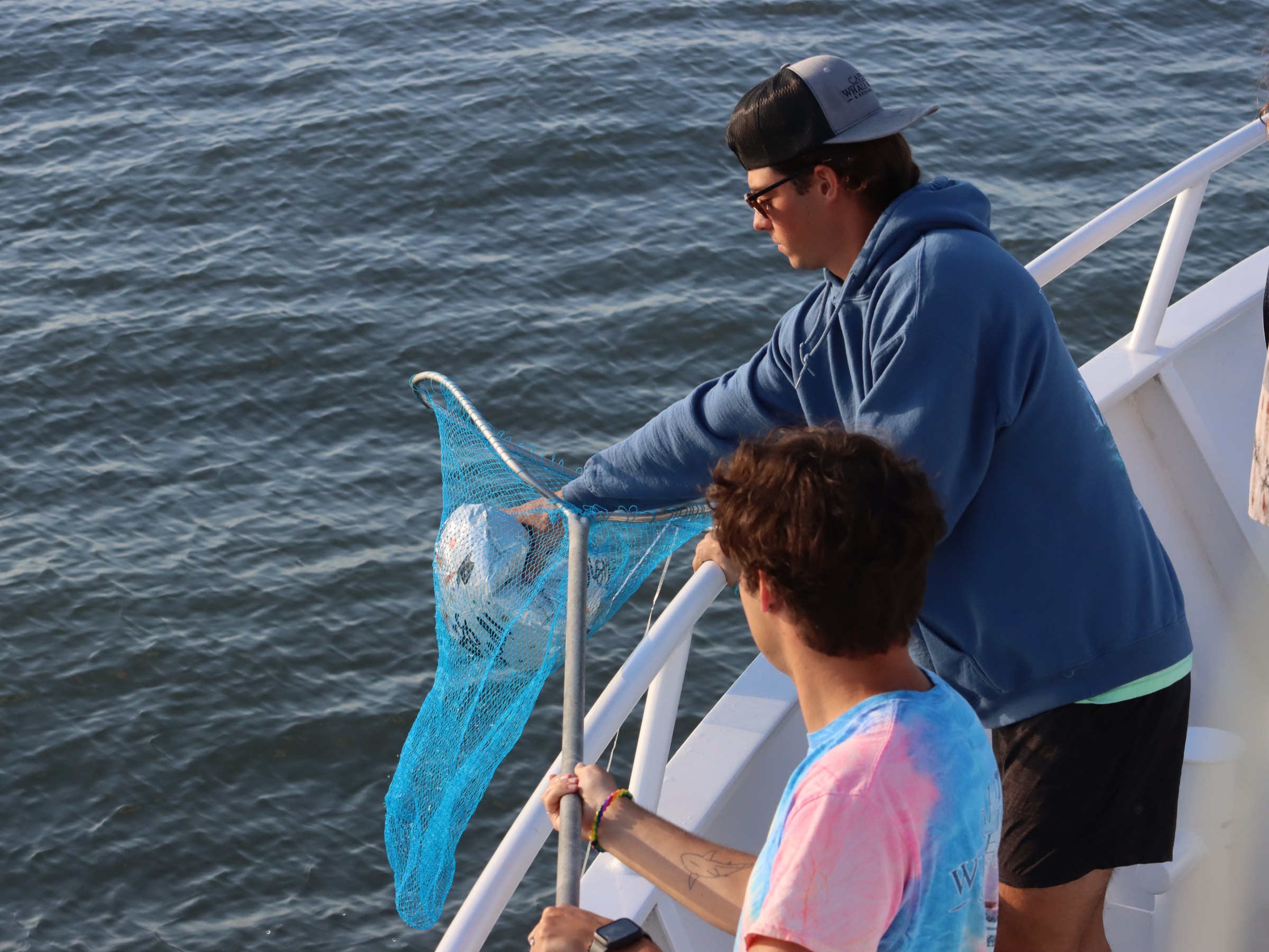 People on a boat near water, one using a net to scoop a floating object.