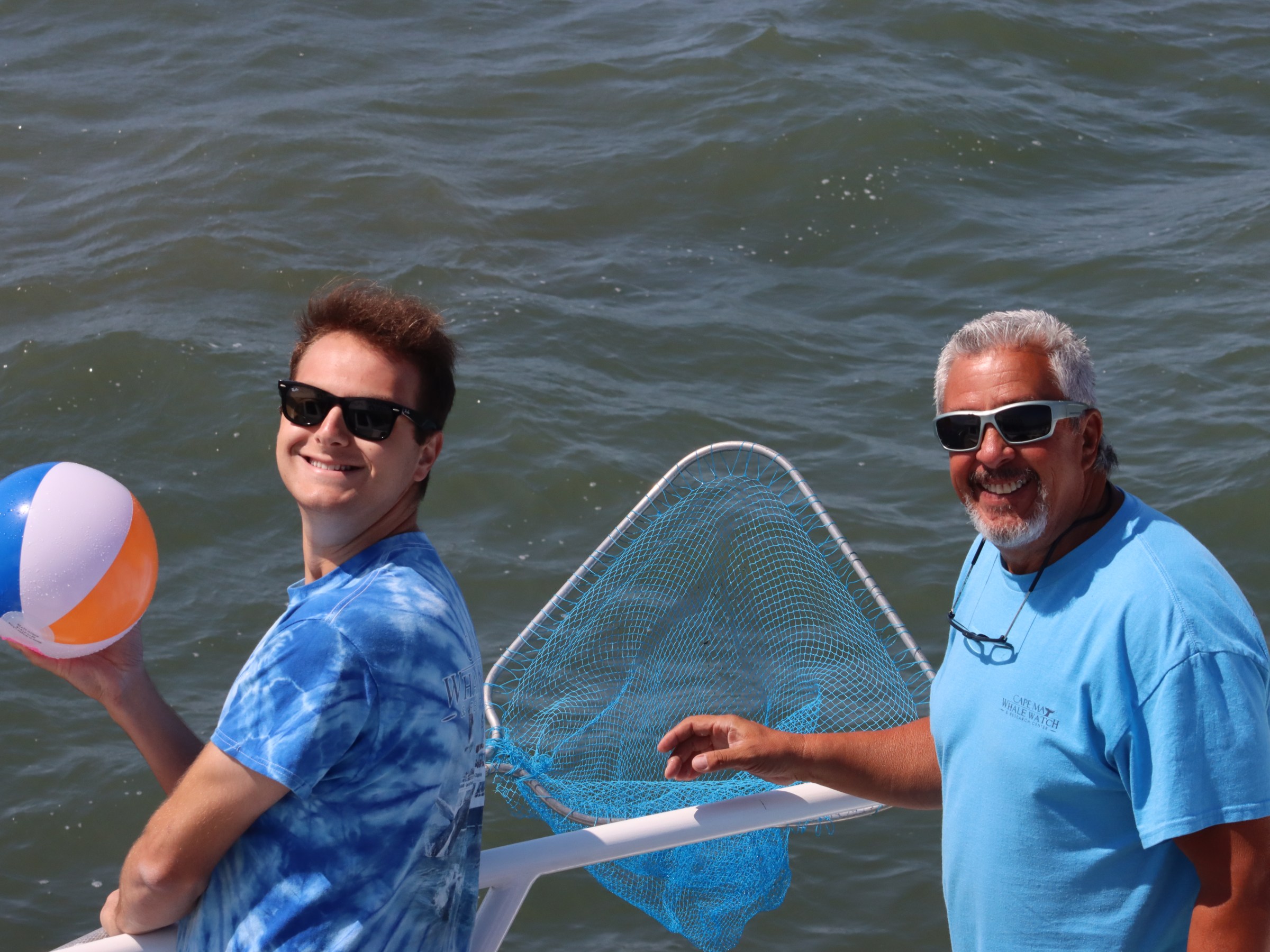 Two men on a boat holding a beach ball and a net, smiling at the camera.