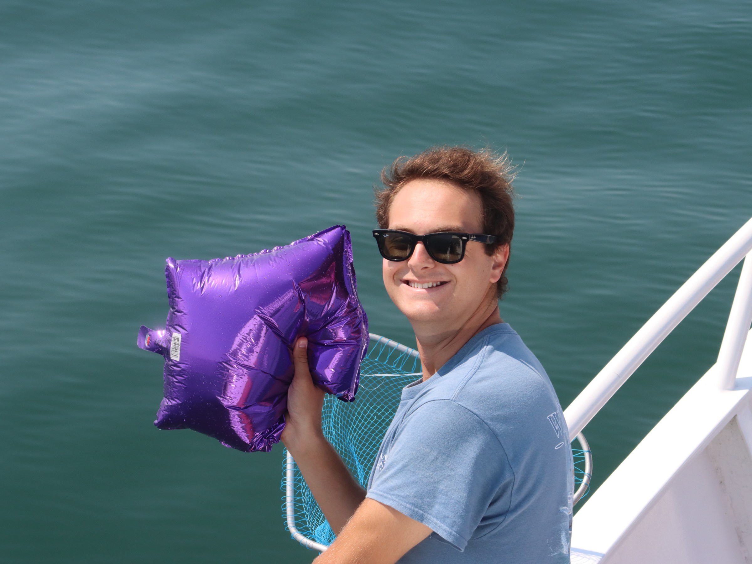 Person in sunglasses holding a purple star-shaped balloon on a boat.