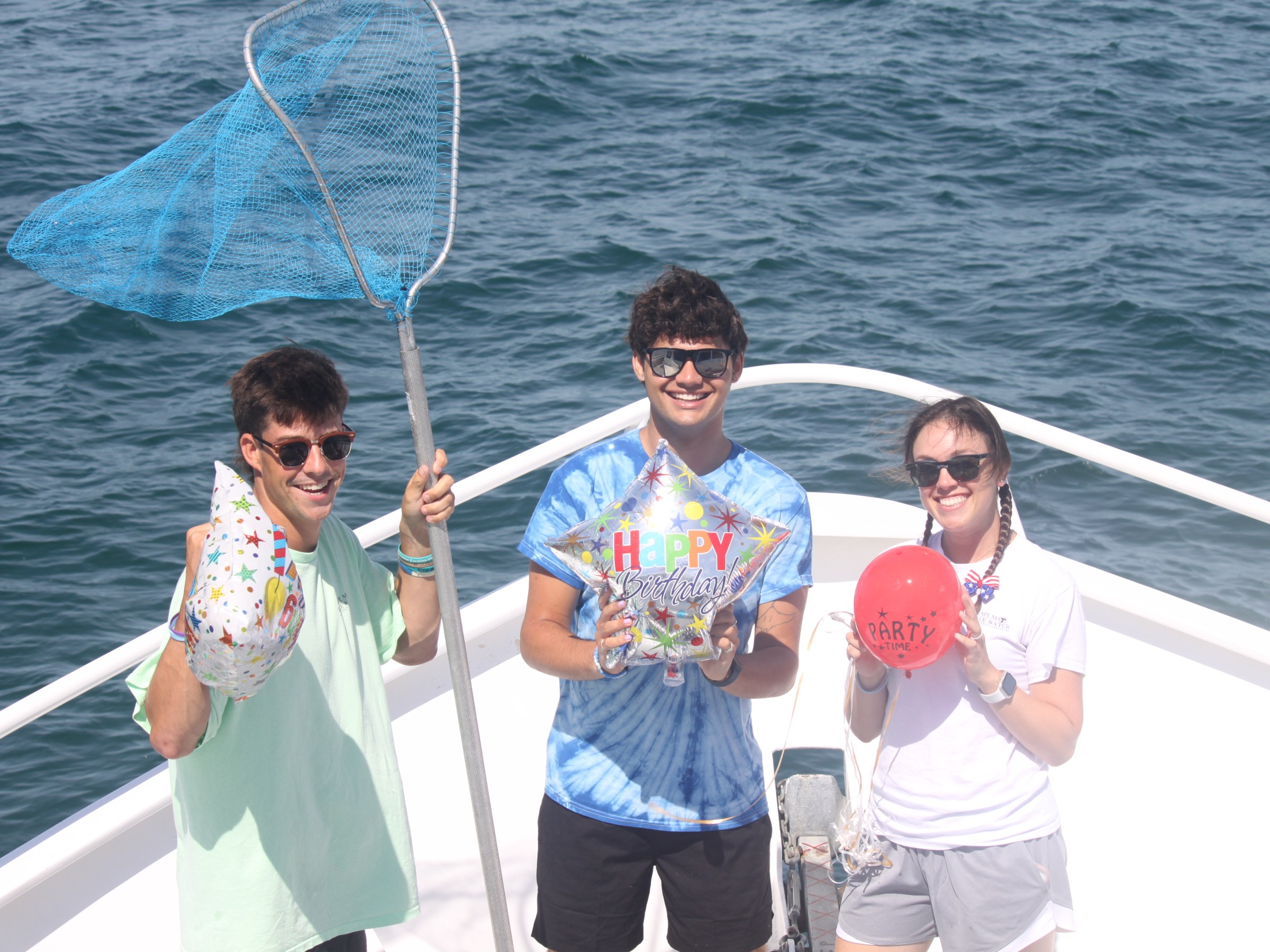 Three people with balloons and a net on a boat, celebrating a birthday.