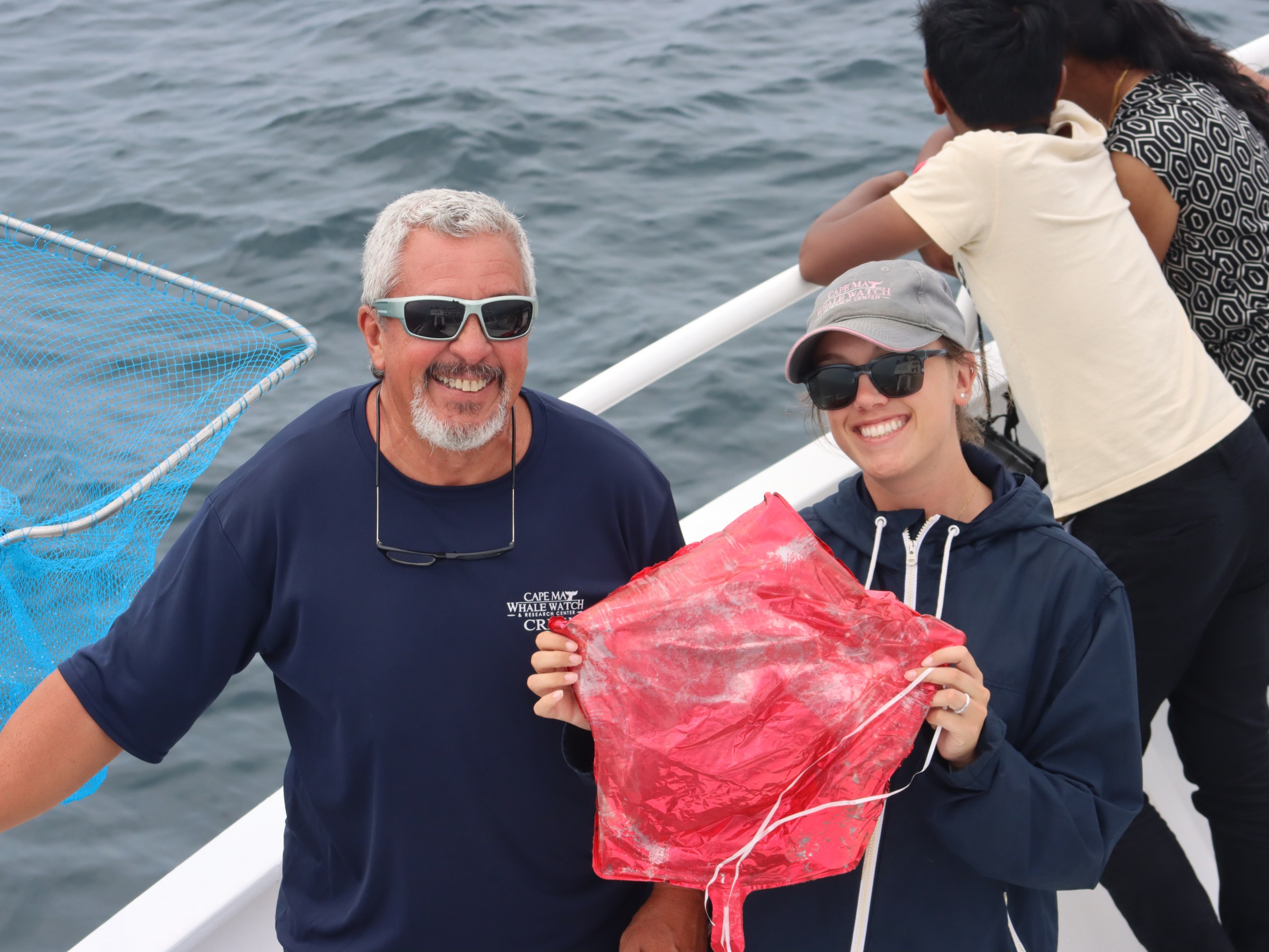 Two people on a boat holding a red object, with another person leaning over the railing.