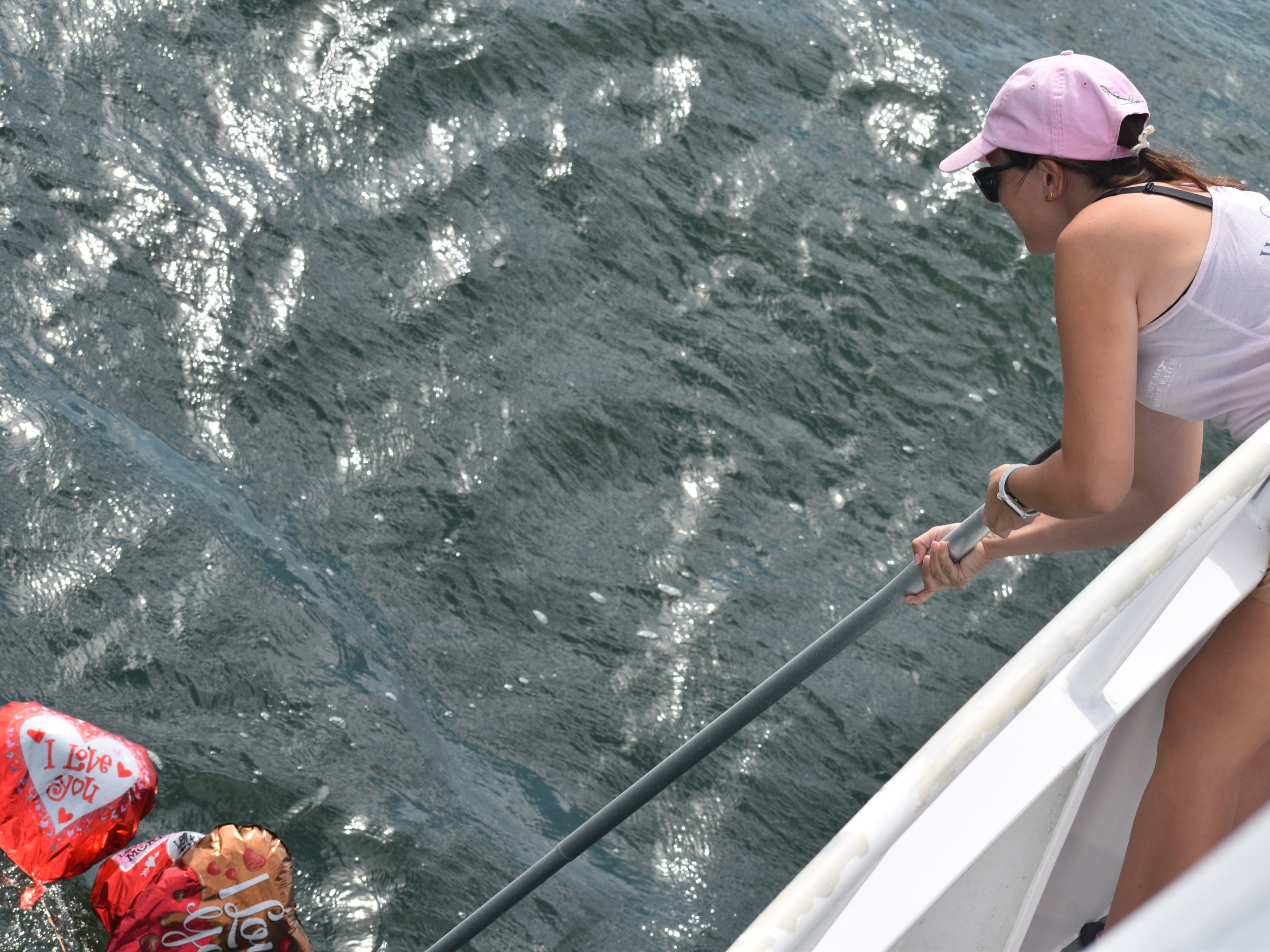 Person on boat using a net to reach heart-shaped balloons floating on water.