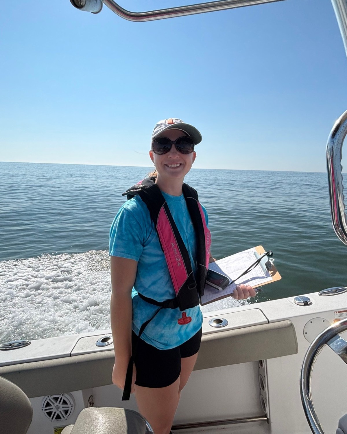 Person wearing a cap and life vest smiles while holding a clipboard on a boat with ocean in the background.