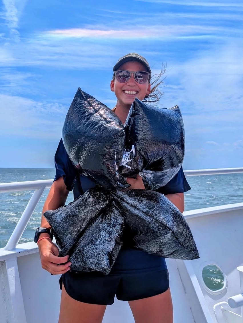 Smiling person holding large black bags on a boat with blue sky and ocean in the background.
